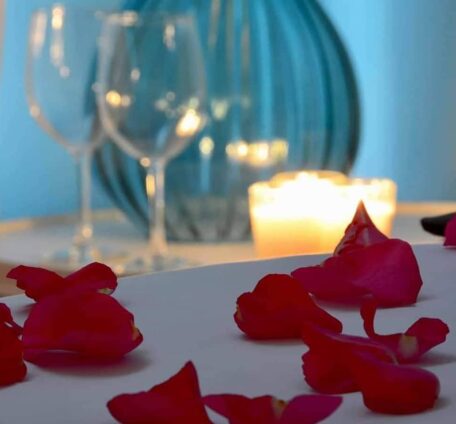 Rose petals strewn on a bed with candles and wine glasses on a table in the background