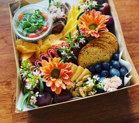Pictured is a box with cheese, fruit, nuts, and spreads, decorated with foliage and flowers.
